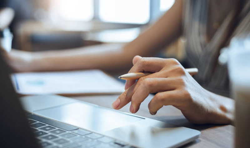 Shadowy image of woman hand poised above laptop keyboard, with a pen resting between her fingers.